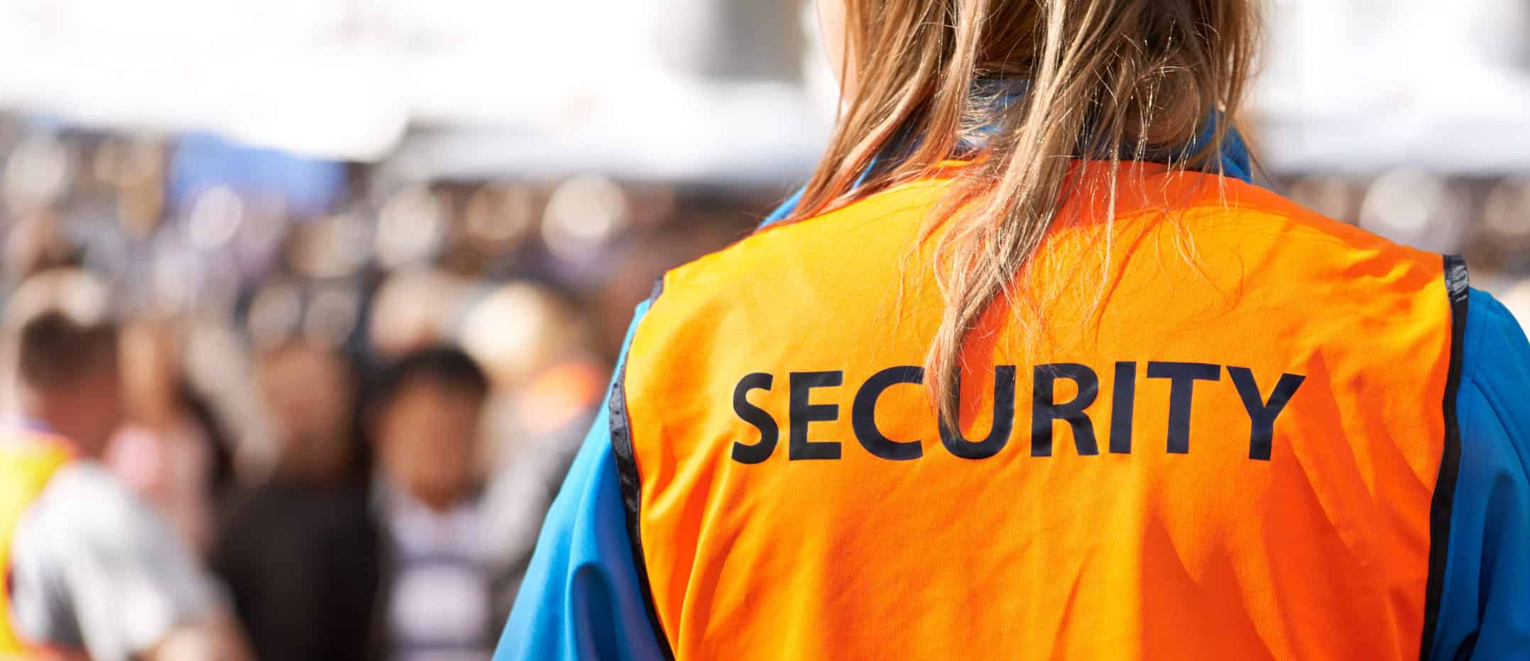 Rear view of a female security guard in orange vest overseeing a crowded outdoor public event for safety.